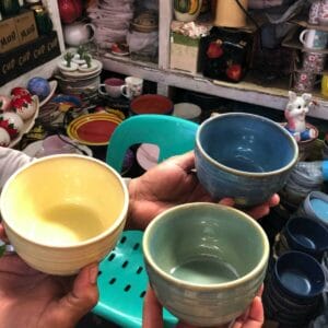 In a store brimming with diverse ceramics and a teal chair, three hands display a set of yellow, green, and blue Soup bowls from Dapitan, highlighting their unique home decor offerings.