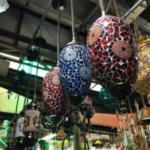 A shop brimming with vibrant Moroccan Lamps - RED/BLUE showcases hanging colorful mosaic lanterns, featuring a stunning array of patterns in blue and red hues.