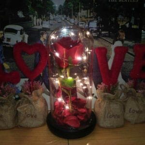 The Big Ben Clock Tower Table Decor, featuring a glass-encased rose with string lights, surrounded by LOVE letters and small bags, makes for an enchanting addition to a wooden surface, ideal for bringing warmth to your home.