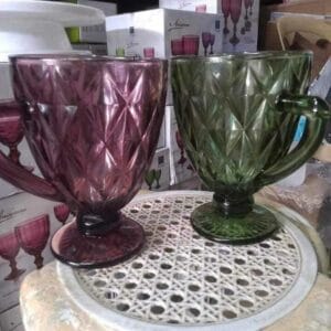 Two Colored Drinking Glasses with a diamond pattern, one in purple and the other in green, rest elegantly on a round white wicker surface, bringing a chic touch to your home decor. Boxes linger subtly in the background, evoking a Dapitan-inspired ambiance.