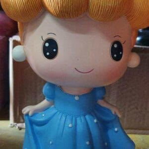 An adorable Assorted Coin-bank featuring a cute doll with a blue dress and orange hair stands charmingly beside a blue hat with a bow, adding a delightful touch of decor to any room.