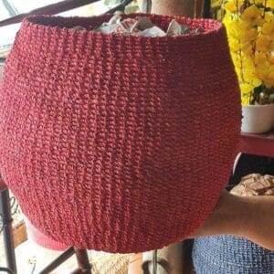 The Soft Abaca Planters, with their red hue and design inspired by Dapitan decor, hang gracefully on a rod, complemented by floral decorations that enhance the background ambiance.