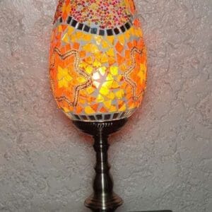 The Table Moroccan Lamp, featuring a mosaic-style design with colorful glass pieces, casts a warm glow on a wooden surface, enhancing your home with elegant decor. This exquisite piece is perfect for creating an inviting atmosphere reminiscent of the vibrant designs found in Dapitan's renowned markets.