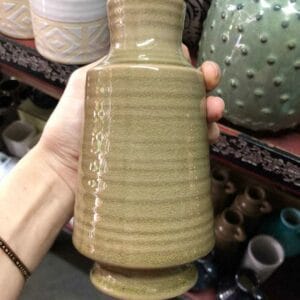 A hand delicately holds the Brown Ceramic Vase with Holder on a store shelf, surrounded by an array of decor treasures, embodying the unique charm of Dapitan.