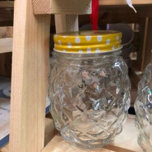 A delightful Pineapple-shaped Glass Jar Mug with a yellow and white polka dot lid sits elegantly on the wooden shelf, adding charm to any home setting and evoking the quaint marketplaces of Dapitan.