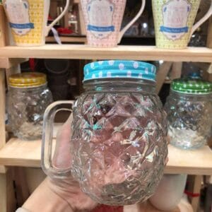 A hand holds a Pineapple-shaped Glass Jar Mug with Lid & Handle, its clear, patterned design and blue polka dot lid perfectly complementing the cozy Dapitan home decor display of mugs in the background.