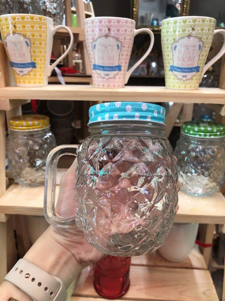 A hand holds a Pineapple-shaped Glass Jar Mug with Lid & Handle, its clear, patterned design and blue polka dot lid perfectly complementing the cozy Dapitan home decor display of mugs in the background.
