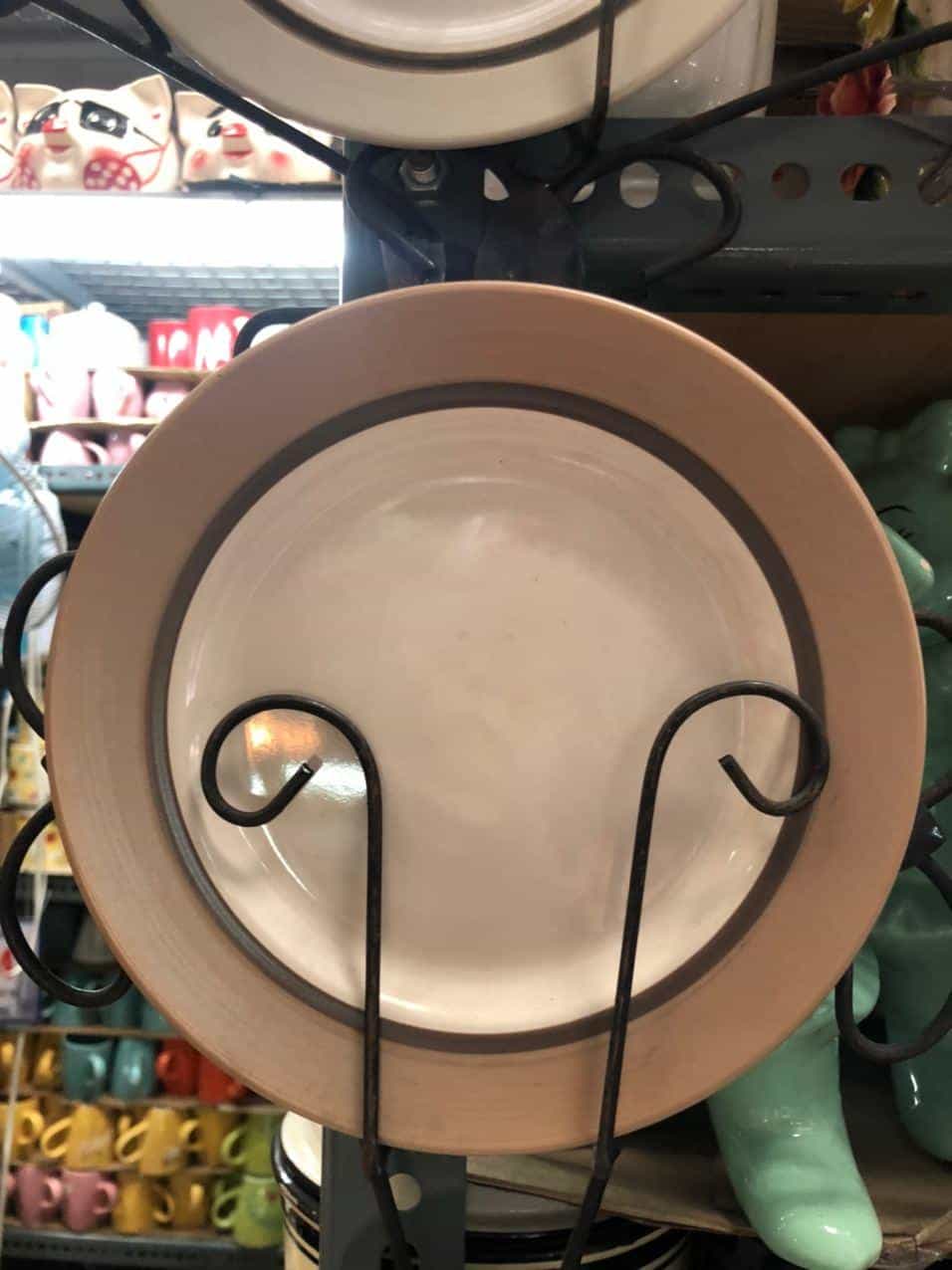 A beige Glossy Round Ceramic Plate with a gray rim rests gracefully on a black metal stand, adding a touch of elegance to your home decor. In the background, shelves display various ceramic items, reminiscent of Dapitan's artisanal charm.
