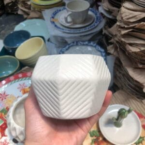 A person delicately holds a White Small Pot amidst an elegant arrangement of stacked plates and cups, embodying the essence of refined home decor. This serene display evokes the charm and allure often found in Dapitan markets.